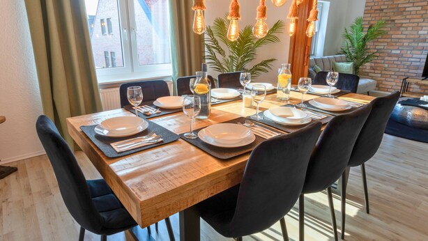 A dining area with a wooden table set for six, black chairs, and hanging light fixtures.