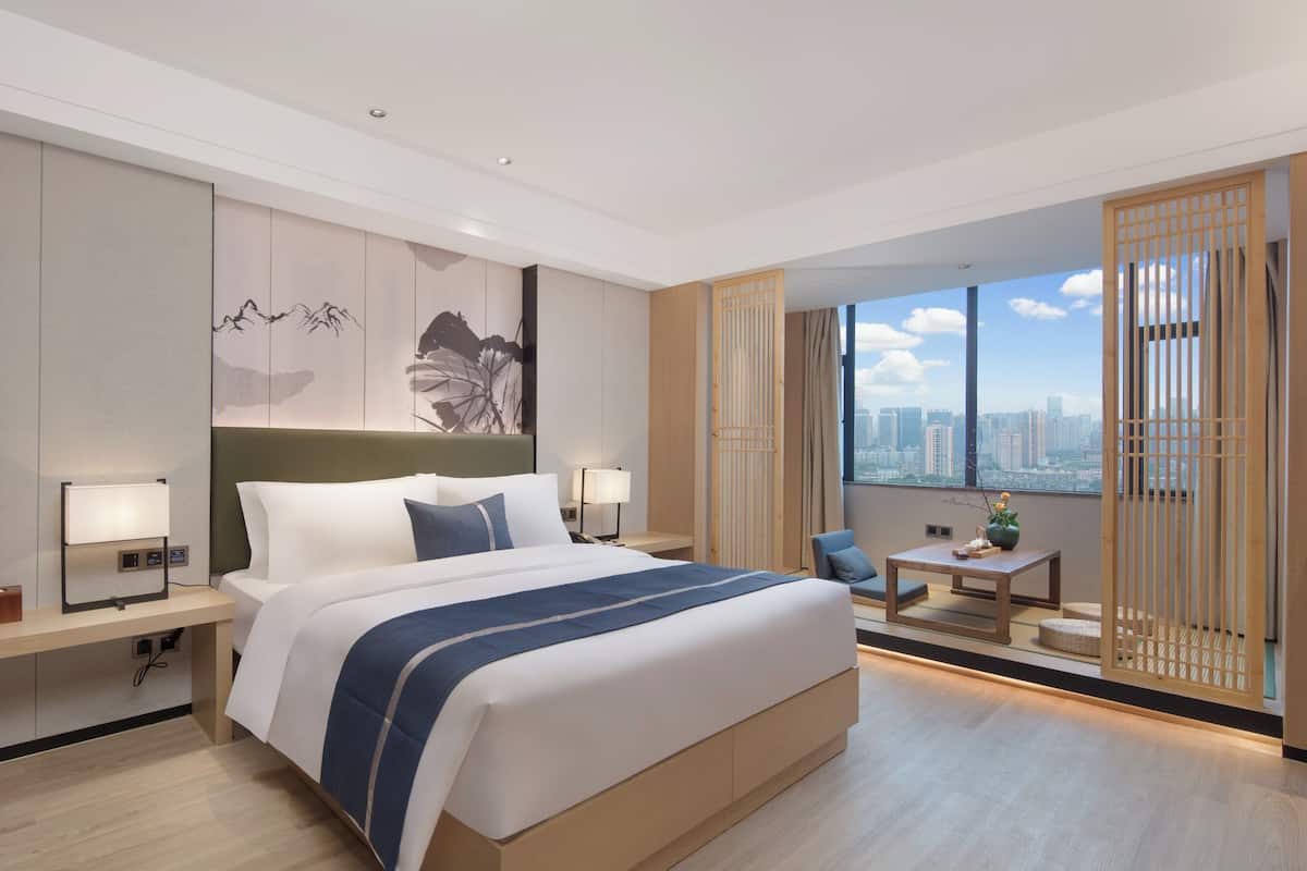 A modern hotel room with a large bed, bedside tables, a desk, and a view of the city.