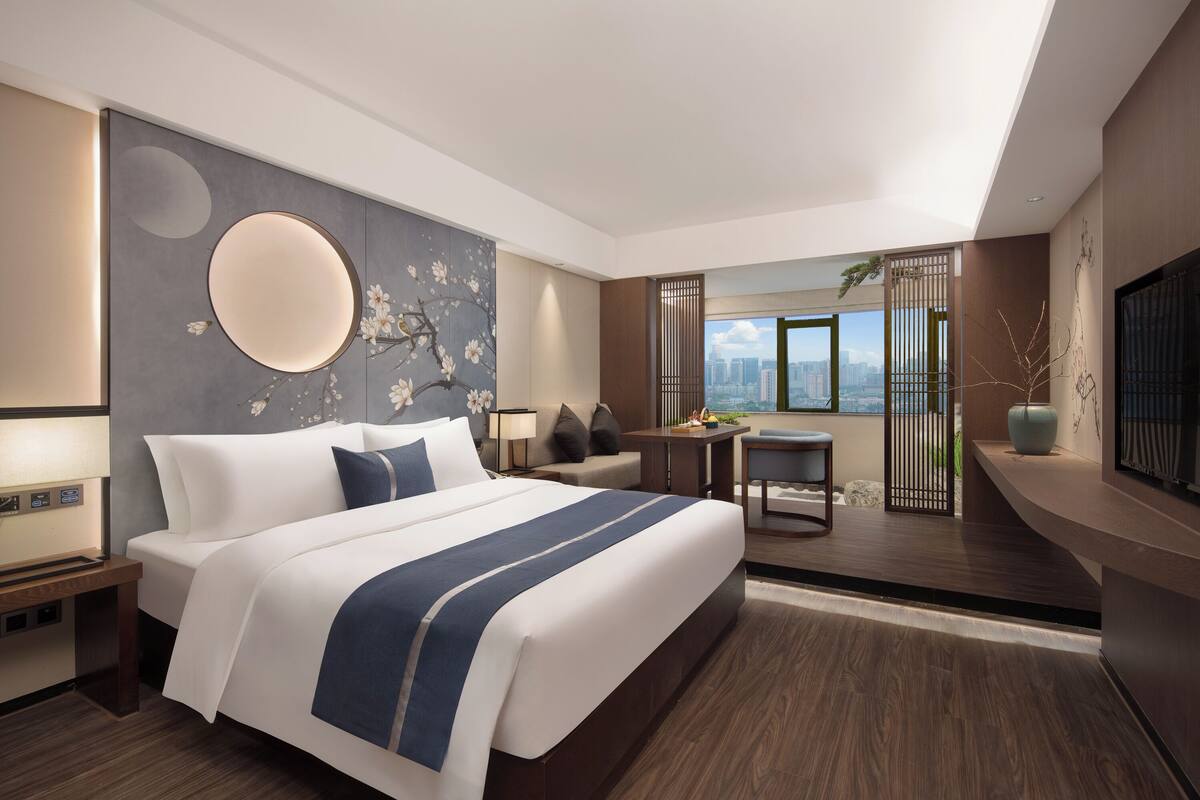 A modern hotel room with a large bed, a desk, a seating area, and a view of the city.