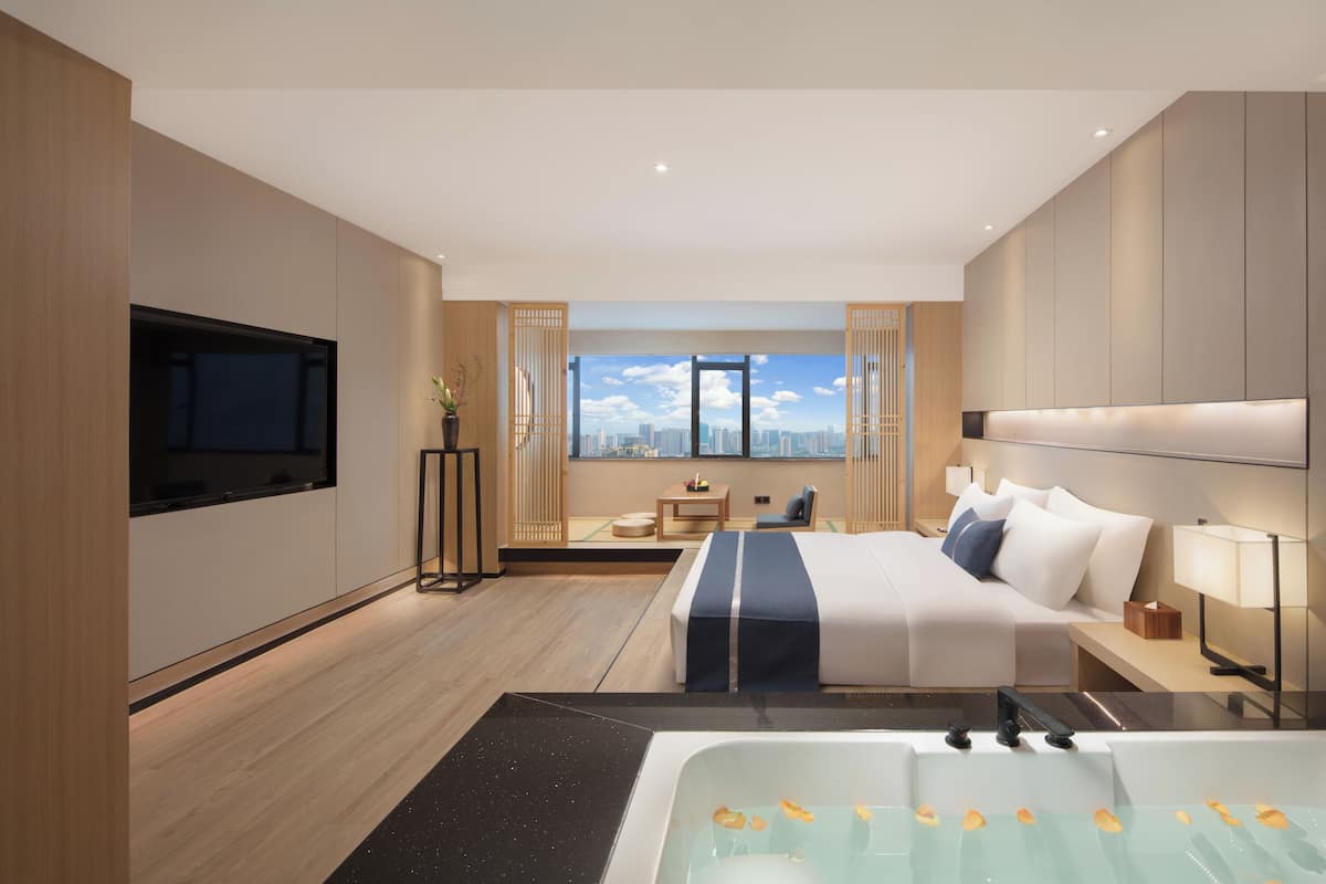 A modern hotel room with a large bed, a sitting area, a view of the city, and a bathtub with rose petals.