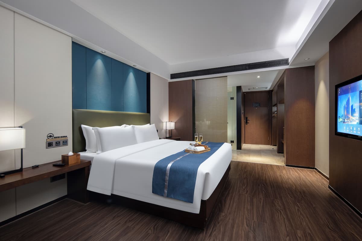 A hotel room with a large bed, a desk with a lamp, a TV on the wall, and a wooden floor.
