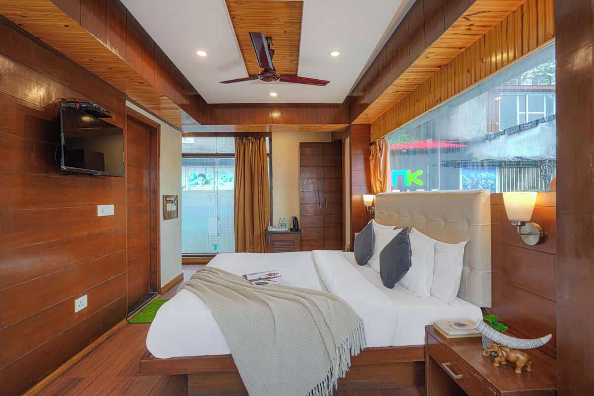 A hotel room with a wooden ceiling, a bed with white bedding, a nightstand with a lamp, and a TV mounted on the wall.