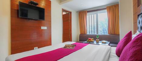 A hotel room with a bed, a mounted TV, a window with curtains, a small table with a plant, and a sofa.