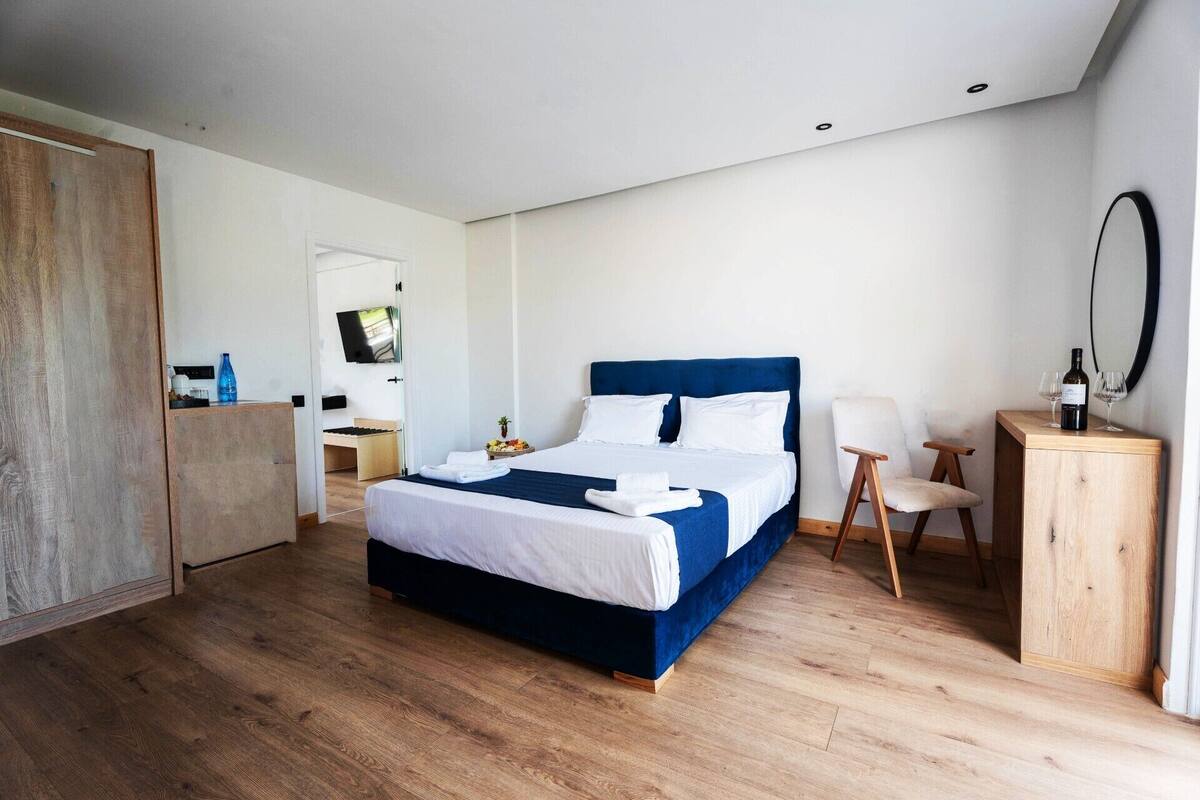 A modern hotel room with a large bed, wooden flooring, a desk, a chair, and a TV.
