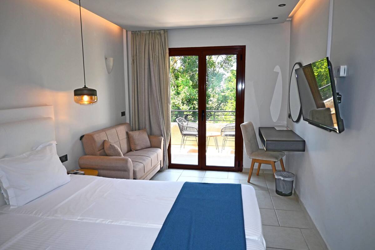 A hotel room with a bed, a sofa, a desk, a chair, a TV, and a balcony with outdoor furniture.
