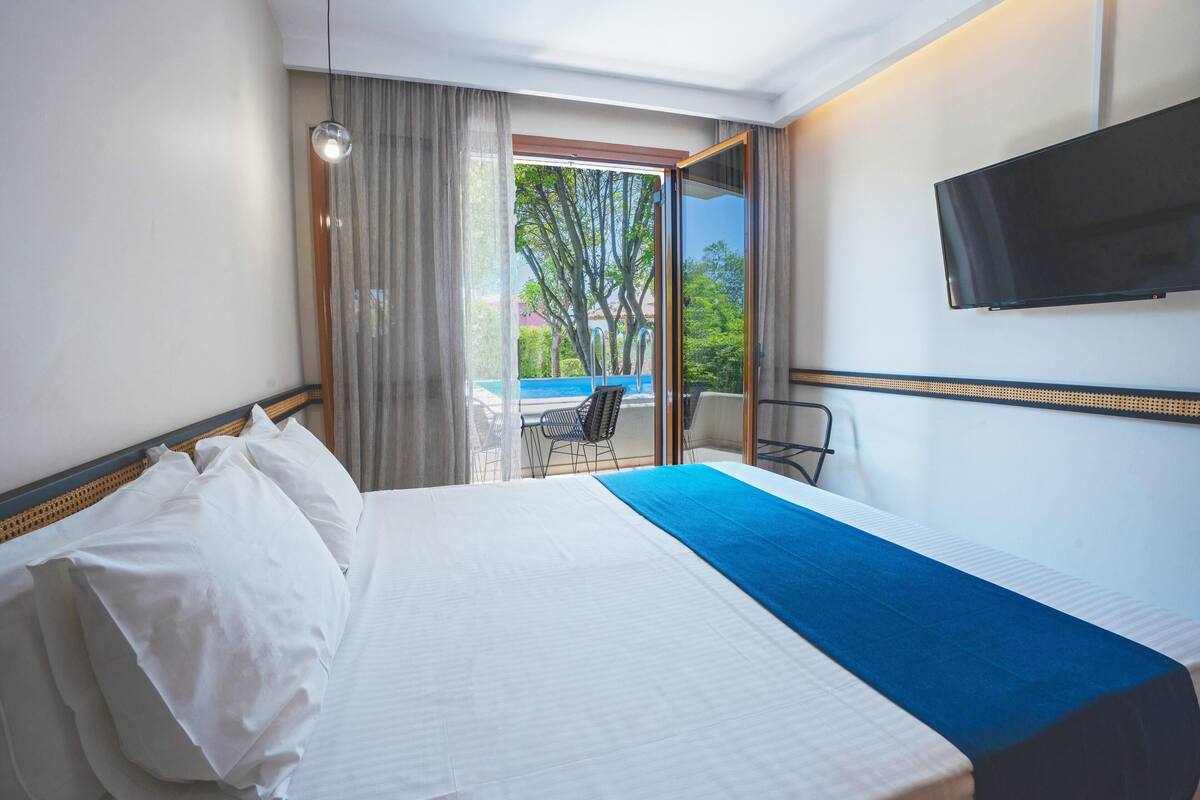 A hotel room with a bed, a TV, a balcony with a chair, and a view of trees and a building.