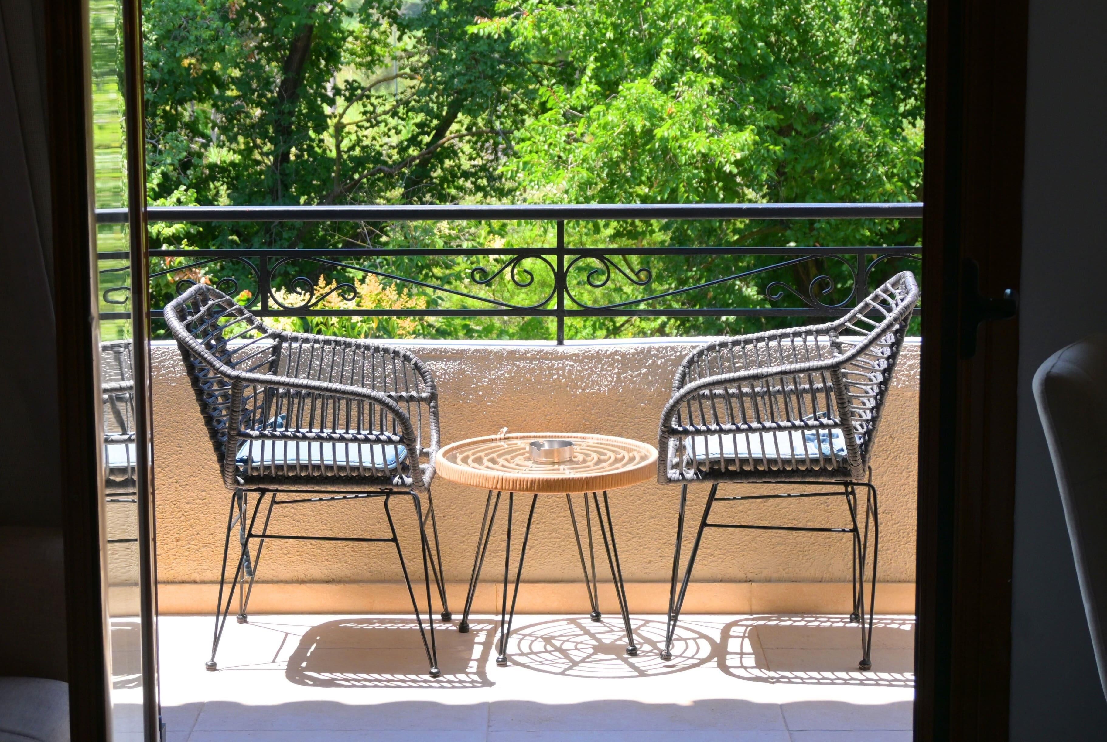 A balcony with two wicker chairs and a small round table, overlooking a lush green area.