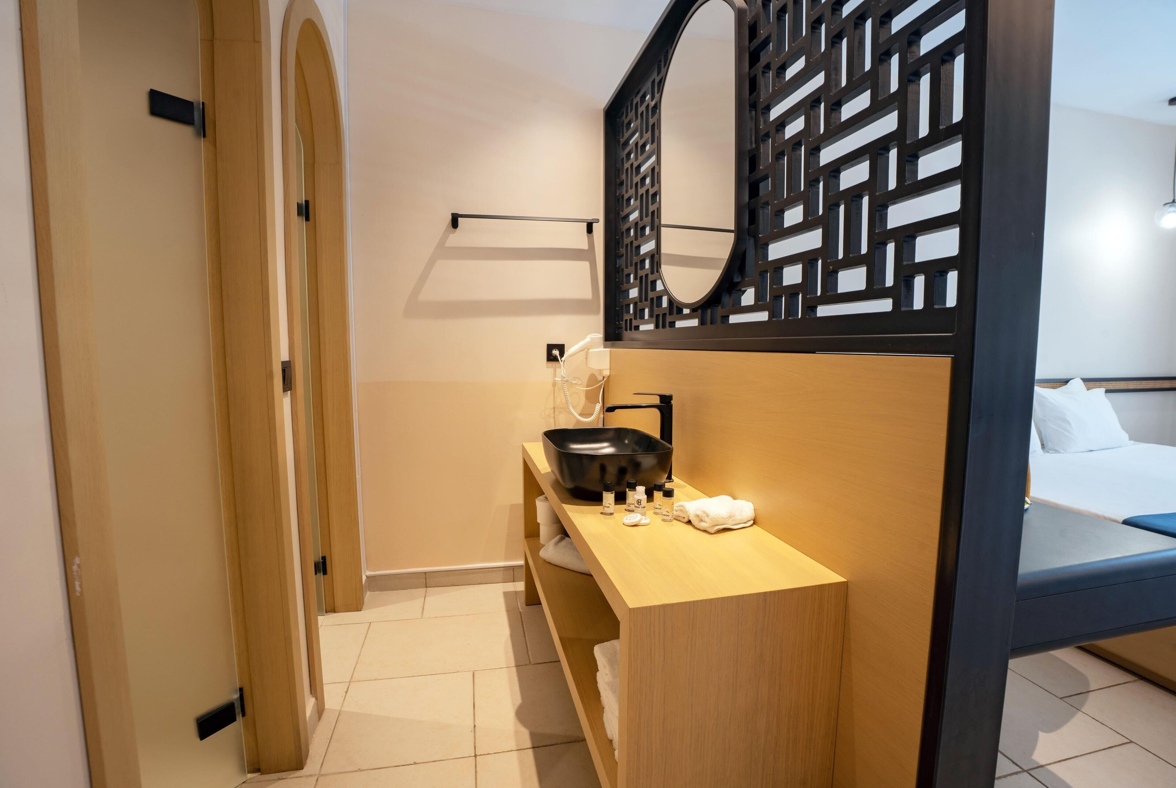 Habitación de hotel con tocador de madera, lavabo negro, toalla y cama con ropa blanca.