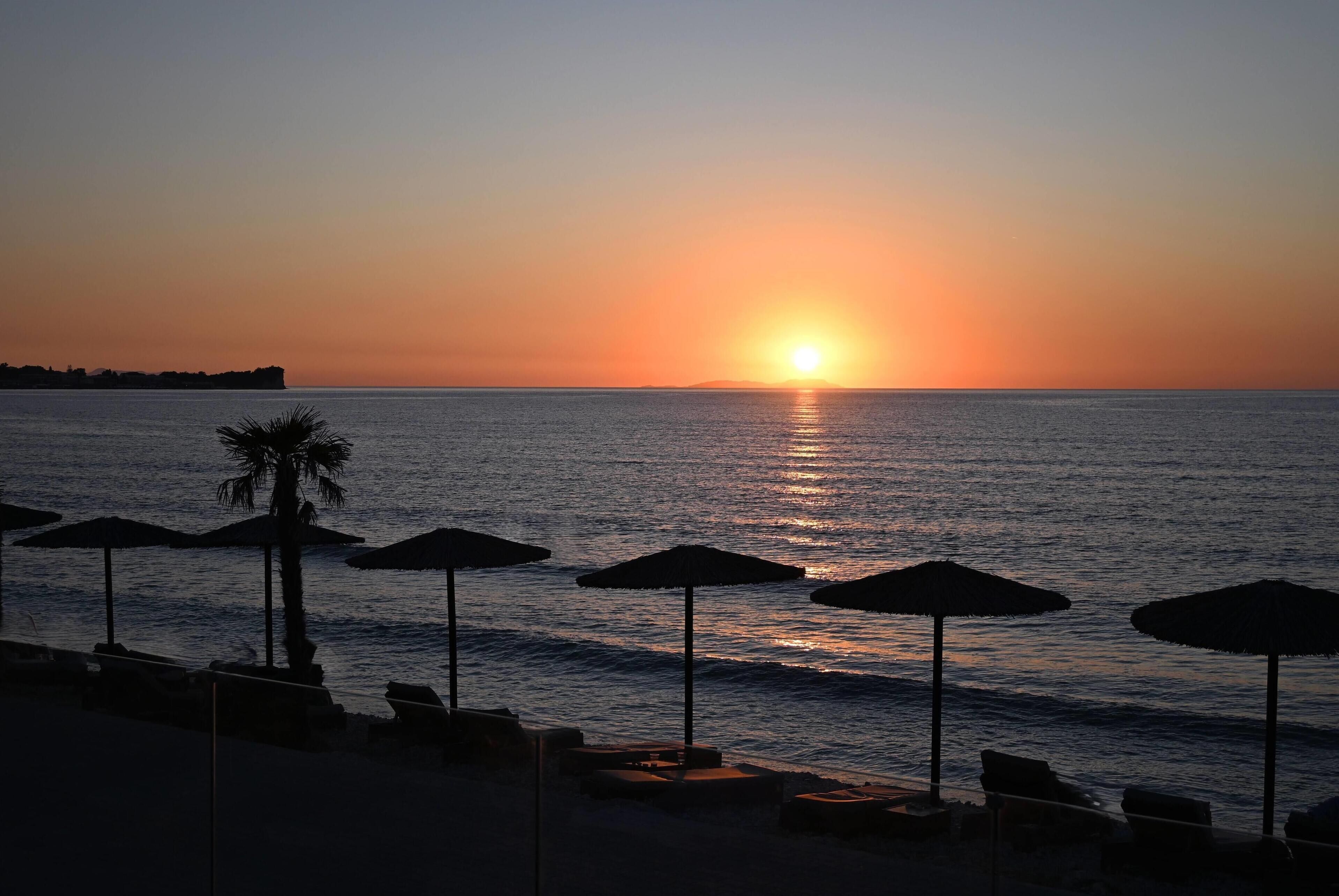 A beach scene with a sunset over the water, umbrellas, and lounge chairs.