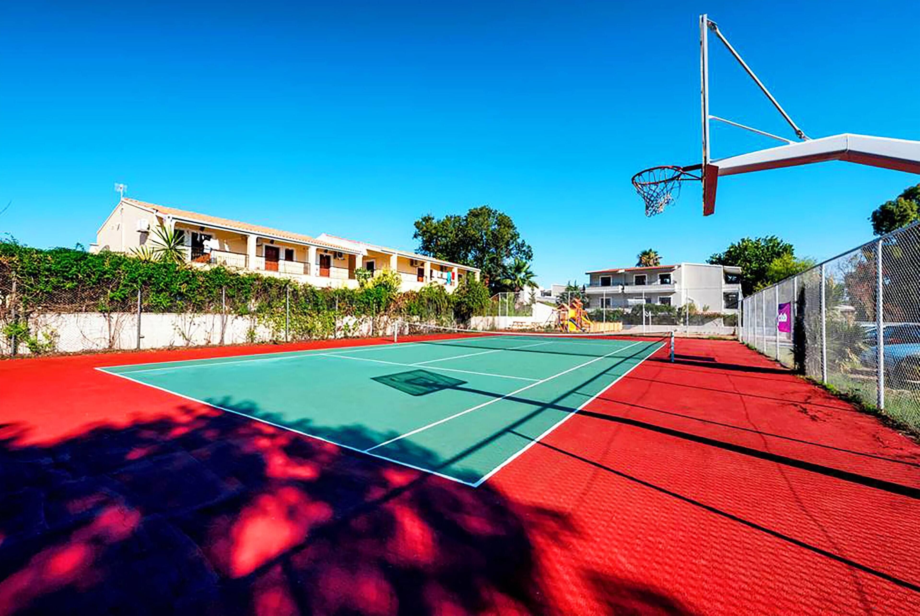 An outdoor basketball court with a red surface, a basketball hoop, and a building in the background.