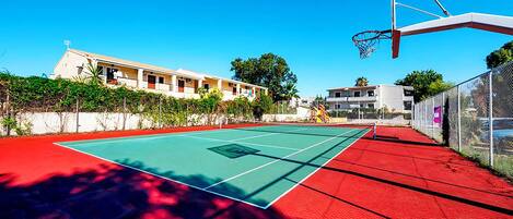 An outdoor basketball court with a red surface, a basketball hoop, and a building in the background.