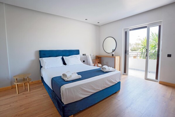 Un dormitorio moderno con una cama grande, una mesita de noche de madera, un espejo redondo y vistas al exterior a través de una puerta corrediza de cristal.