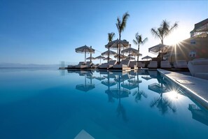 Una piscina con sillas de descanso y sombrillas, palmeras y un cielo despejado.