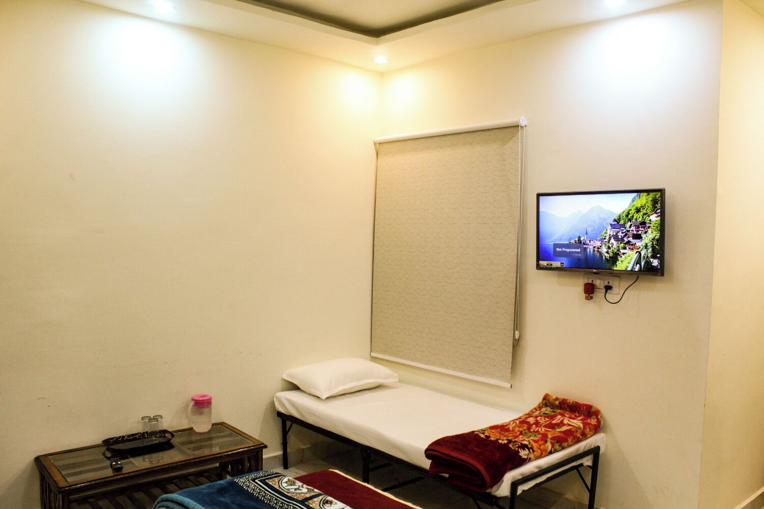 Una habitación pequeña y sencilla con una cama individual, un televisor fijado a la pared y una mesita con un control remoto y una botella.