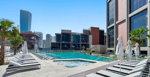 A rooftop pool area with lounge chairs and umbrellas, surrounded by modern buildings and a clear sky.