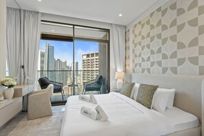 Et moderne hotelværelse med en stor seng, to stole, et skrivebord og udsigt til byens skyline gennem gulv til loft-vinduer.