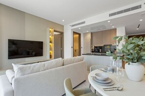Een moderne woonkamer met een bank, een eettafel met borden en glazen, een aan de muur bevestigde televisie, en een zichtbare keuken door een open doorgang.