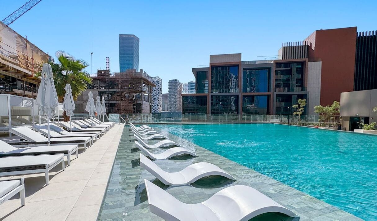 A rooftop pool with sun loungers and umbrellas, surrounded by modern buildings and a clear sky.