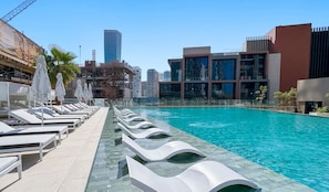 A rooftop pool with sun loungers and umbrellas, surrounded by modern buildings and a clear sky.
