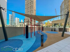 An outdoor fitness area with exercise equipment, a shaded seating area, and a view of the city skyline.