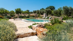 A pool area with a clear blue sky, surrounded by greenery and rocks. - Trulli Pietra Rossa With Pool by Wonderful Italy (Ostuni)
