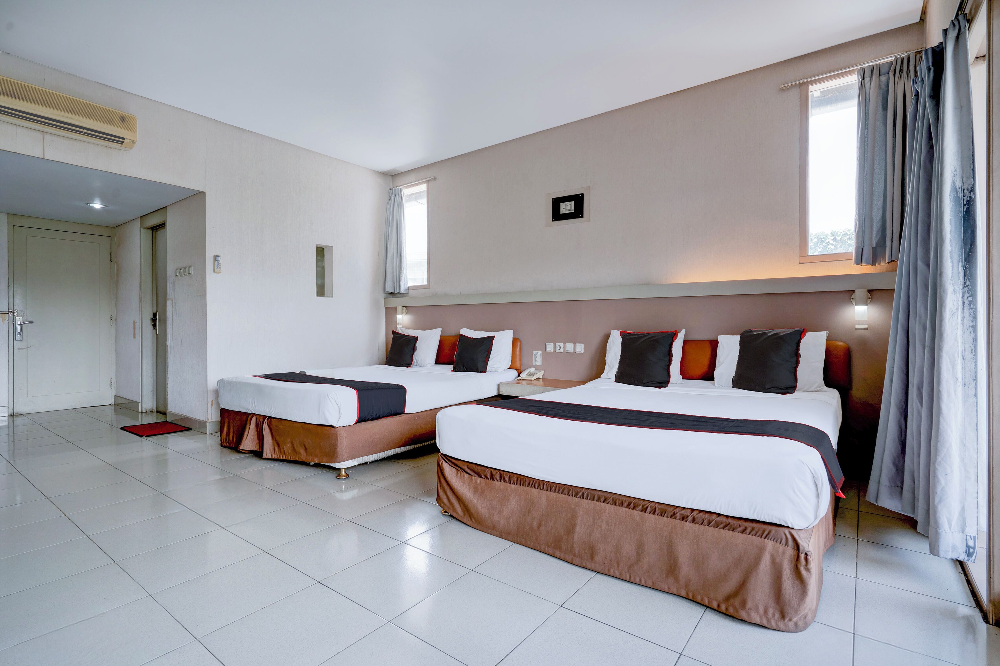 A hotel room with two beds, a red mat, a window with curtains, and a door.