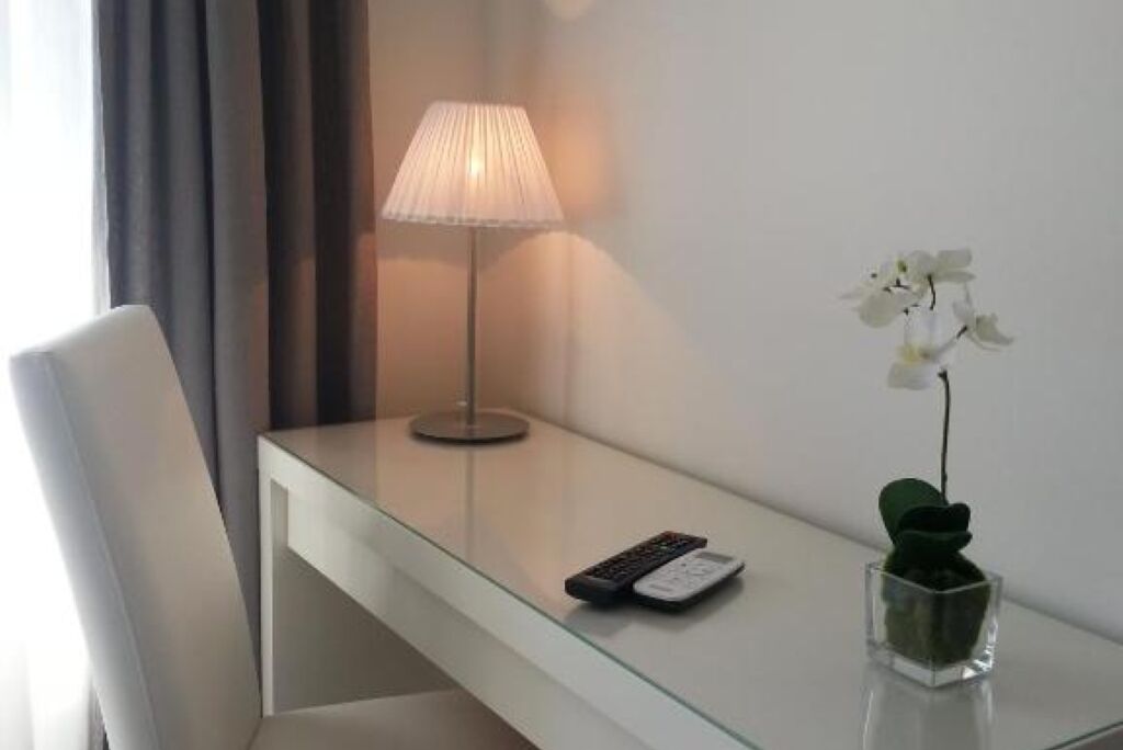 Habitación de hotel con un escritorio de cristal, un control remoto, una lámpara y una pequeña planta de orquídea.
