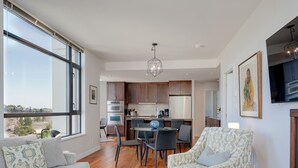 A modern living room with a sofa, armchair, and dining area. There is a kitchen in the background with a large window.