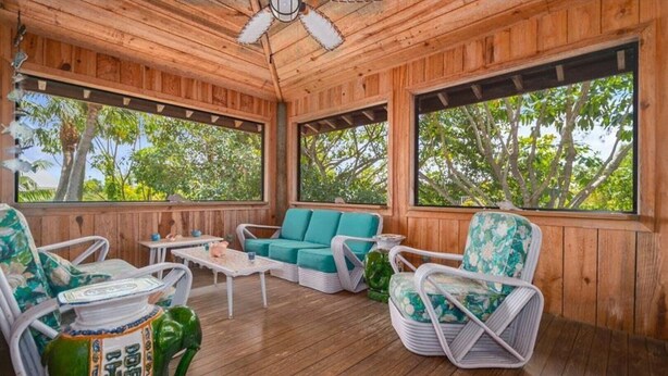 A wooden deck with wicker furniture, a ceiling fan, and large windows overlooking a forested area.