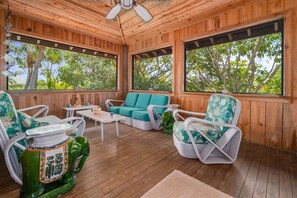 A wooden deck with wicker furniture, a ceiling fan, and large windows overlooking a forested area.