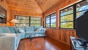 A cozy living room with a sectional sofa, a coffee table, and large windows offering views of greenery.
