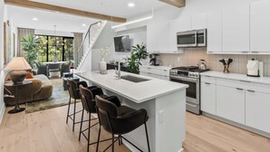 A modern kitchen with white cabinetry, a central island with bar stools, a stove, microwave, and a large window offering a view of greenery. - Olive Stone Modern Penthouse Near Mercedes Benz (Atlanta)