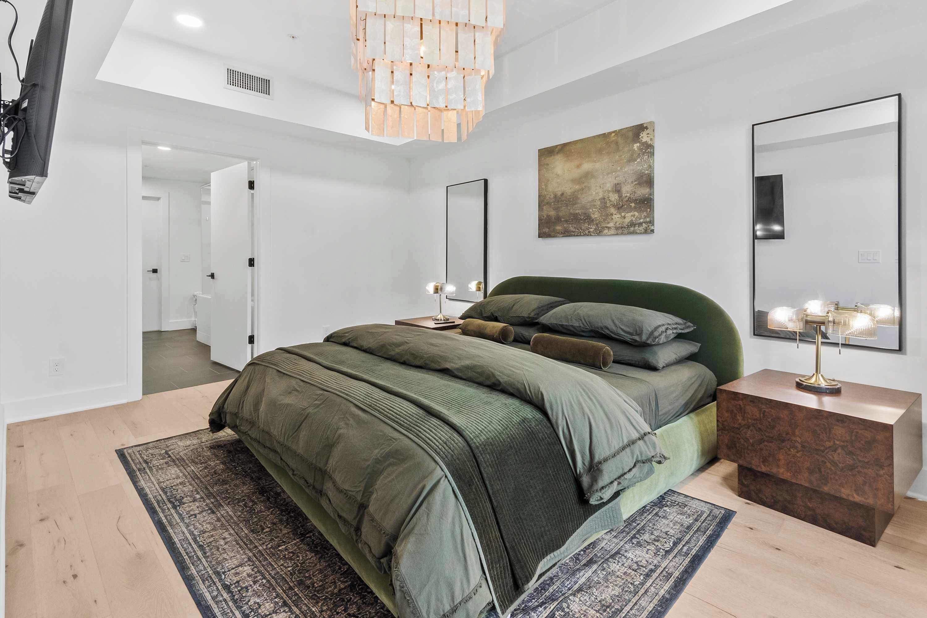 Ett modernt sovrum med en grön klädd säng, en stor spegel och en kristallkrona.