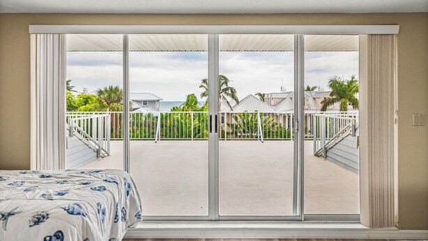 A room with a large sliding glass door leading to a balcony with a white railing, a view of a residential area with palm trees, and a bed with a patterned bedspread. - 2740 Shore Ln - 1 block to the peach - Pet Friendly!  Large Home (Boca Grande)