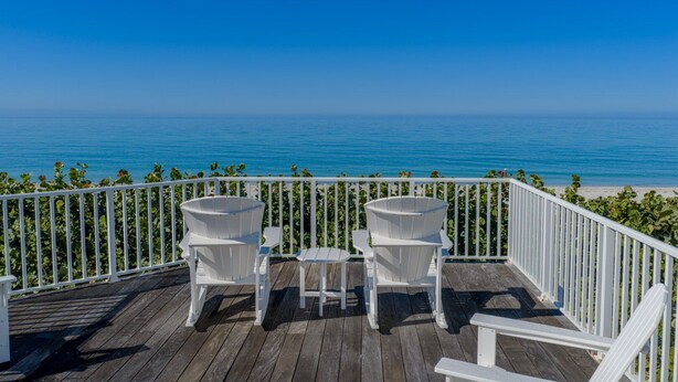 A wooden deck with two white Adirondack chairs and a small table, overlooking a beach and the ocean.