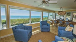 A spacious living room with large windows offering a view of the ocean, a ceiling fan, and a dining area with wooden furniture.