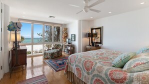 A bedroom with a large window, a ceiling fan, a bed with patterned bedding, a chair, a small table, and a bookshelf. - 2973 Shore Ln - 4 Bedroom - Beachfront Home (Boca Grande)