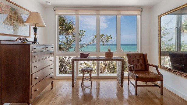 A room with a wooden dresser, a table with a woven stool, a chair, and large windows offering a view of the ocean and trees. - 2973 Shore Ln - 4 Bedroom - Beachfront Home (Boca Grande)
