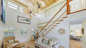 A spacious living area with a high ceiling, a wooden staircase, and a large window with shutters.