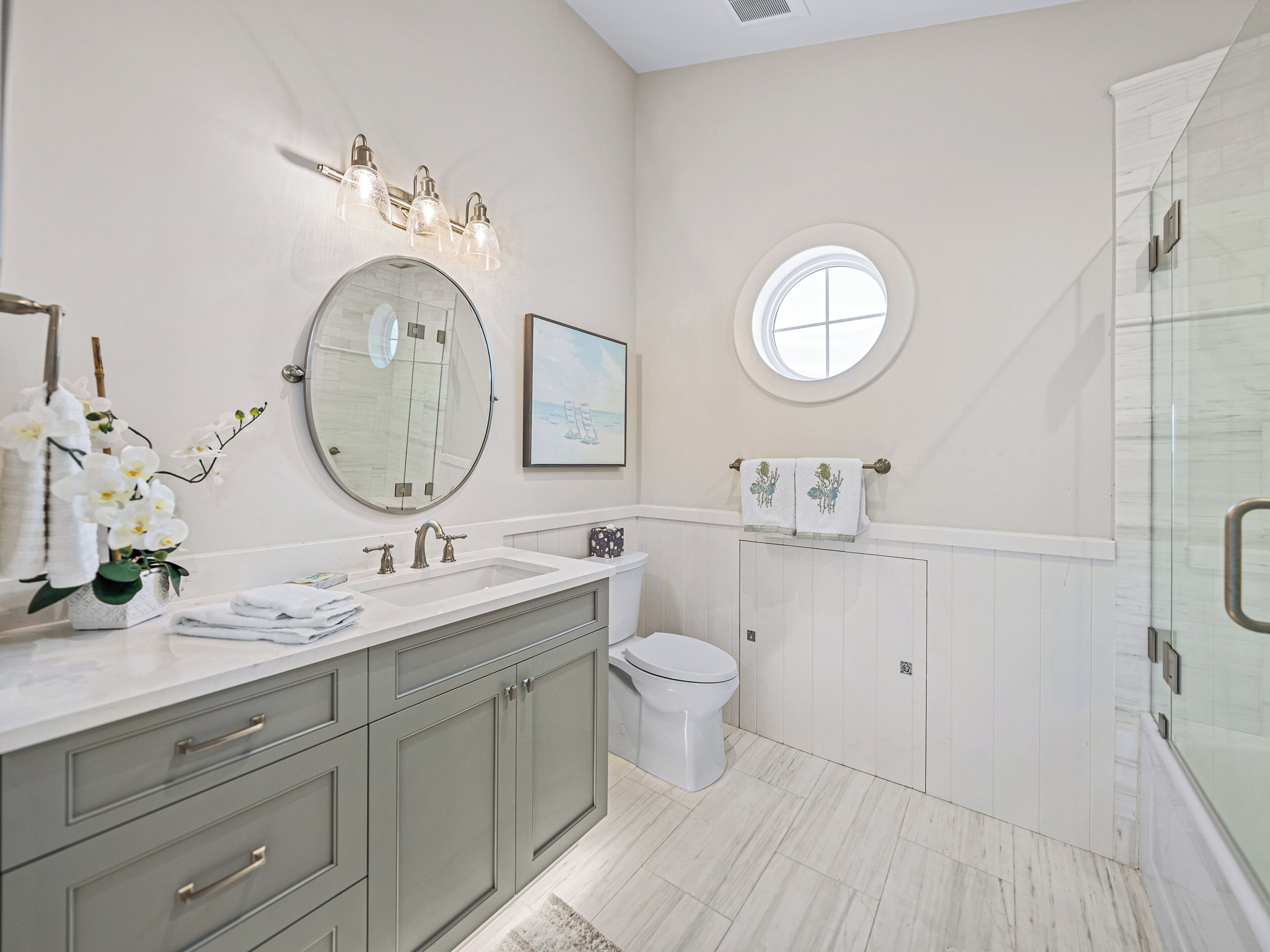 A modern bathroom with a vanity, sink, and toilet. There is a round mirror above the sink, a window with a view, and a shower area.