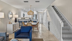 A modern living room with a blue sofa, a dining area with wicker chairs, a kitchen with white cabinets, and a staircase with patterned carpet. - Sundown 03:  2-bed, 2-bath townhome in Boca Grande stunning Gulf front views, a relaxing getaway. (Boca Grande)