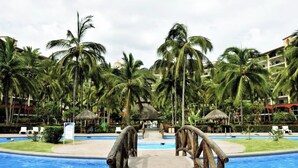 A resort pool area with palm trees, a thatched-roof hut, and a view of hotel buildings in the background.