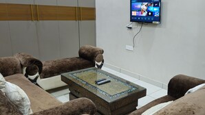 A living room with a brown sofa set, a glass coffee table with a remote control, and a mounted TV displaying various channels. - Royal Stay Inn (Moradabad)