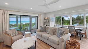 A spacious living room with a large sofa, a round coffee table, and a dining area with chairs. A balcony with a view of the ocean is visible through large windows. - Luxury Seagrape 01, 3-bedroom, 3-bathroom townhome in Boca Grande with stunning Gulf views. (Boca Grande)