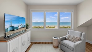 A room with a large window overlooking a beach, a television on a cabinet, and a patterned side table. - Luxury Seagrape 01, 3-bedroom, 3-bathroom townhome in Boca Grande with stunning Gulf views. (Boca Grande)