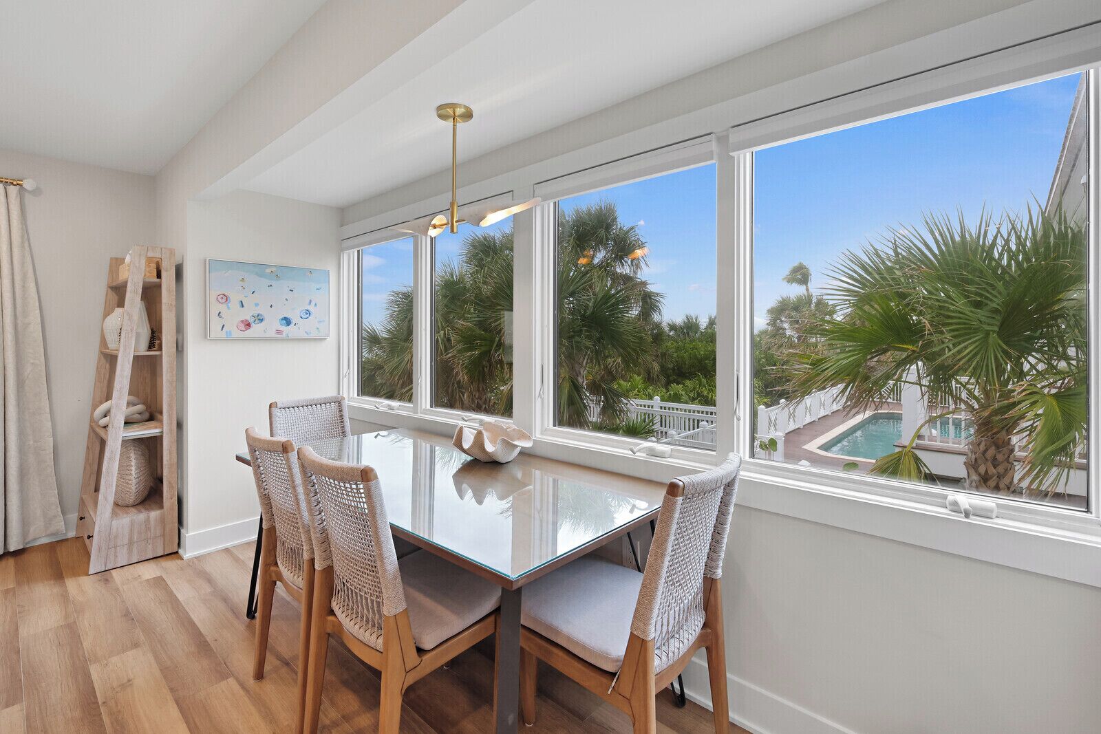 A dining area with a glass table and wicker chairs, large windows with a view of palm trees and a pool, and a wooden ladder against the wall.