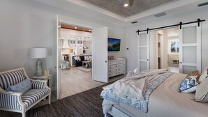 A spacious bedroom with a large bed, a seating area with a chair, and a view into the dining area.