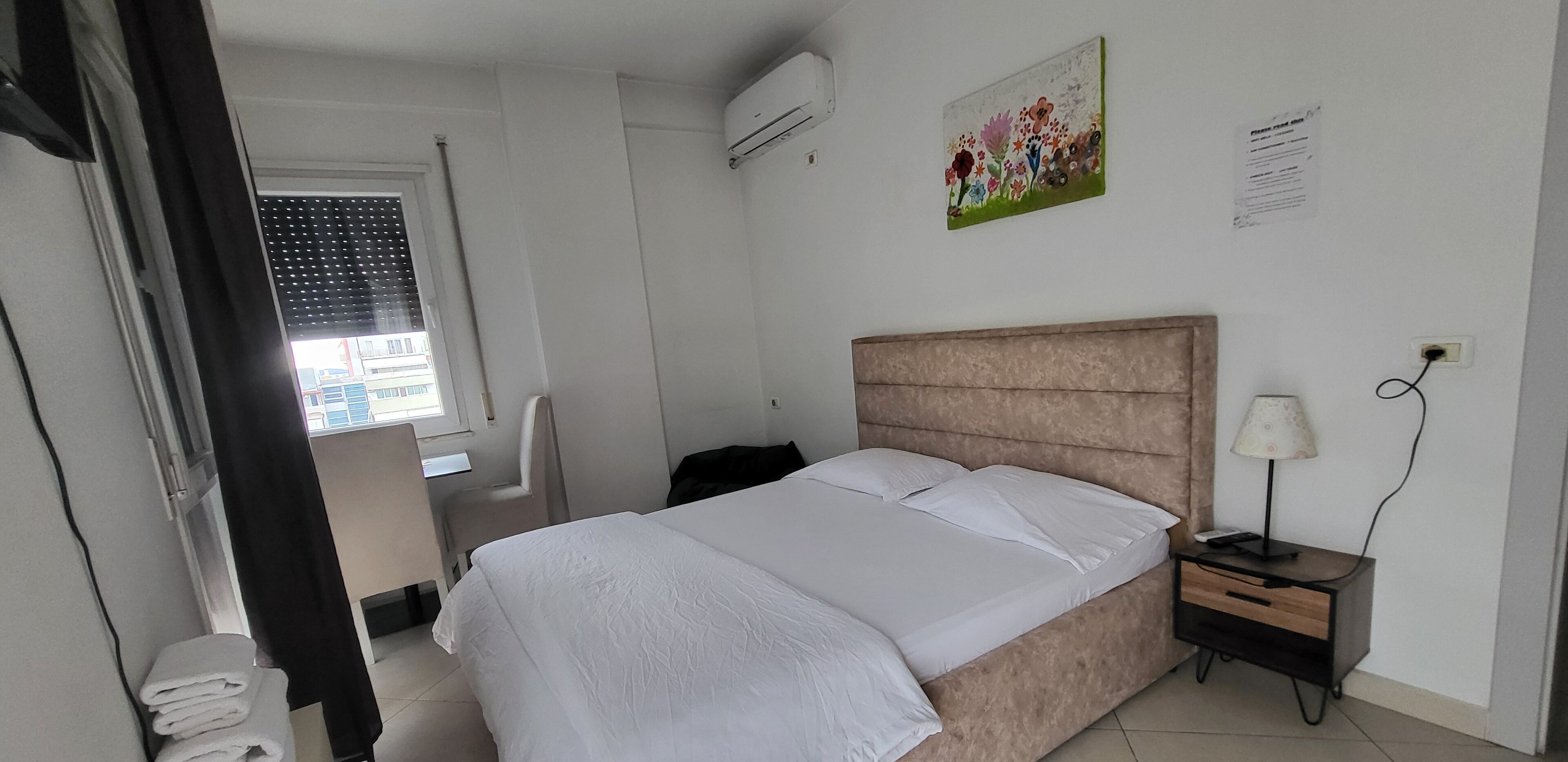Una habitación de hotel con cama, escritorio, silla, mesita de noche y ventana con persianas.
