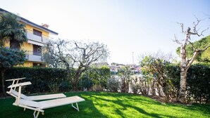 A garden with two wooden lounge chairs, a yellow building with balconies, and various trees and shrubs. - Villaggio dei Fiori - App Primula (Sirmione)