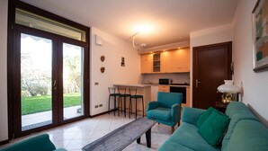 A living room with a sofa, a coffee table, and a kitchen area with a dining table and chairs. - Villaggio dei Fiori - App Primula (Sirmione)